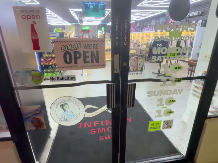 Glass front doors with a Come In We're Open sign, store hours lettering and the infinity doormat