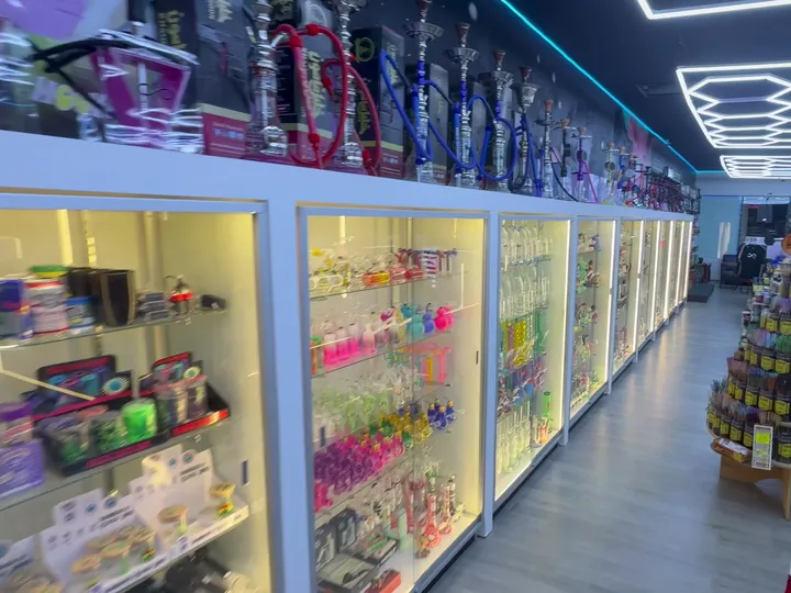 Aisle of lit glass cases with colorful glass pieces and hookahs above, beside a sock rack